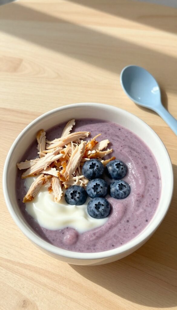 A dog-safe chicken and blueberry smoothie bowl on a wooden table, topped with blueberries and shredded chicken, with a dog bowl and spoon beside it.